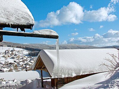 Bilderbuchwinter in Sankt Englmar. Verschneite Dächer mit hängenden Eiszapfen. Im Hintergrund der zum Hirschenstein führende Winterwald.