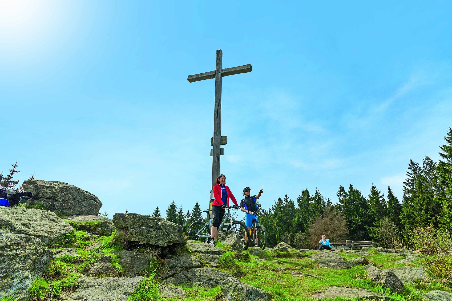 Ein junges Paar steht mit ihren Mountainbikes direkt am Gipfelkreuz des Pröllers. Der junge Mann zeigt in die Ferne. In der Nähe sitzt eine Frau auf einer Bank und genießt Wald und Sicht. Über allem ein blauer Himmel.