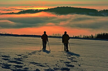 Zwei Schneeschuhwanderer auf freier Fläche. Sie gehen auf glitzerndem Schnee auf ein atemberaubendes Farbenspiel aus Nebel und orangerotem Sonnenlicht zu.