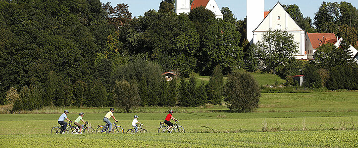 Vier Erwachsene und ein Kind radeln auf einem, durch eine Wiese führenenden Weg. Im Hintergrund ragen zwischen Bäumen die beiden Kirchtürme der Wallfahrts- und Kreuzkirche Haindling  in den blauen Himmel.