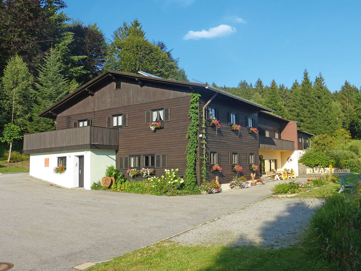 Das Gruppenhaus namens BayerWaldAktivZentrum liegt idyllisch im Wald. Darüber strahlt der blaue Sommerhimmel.