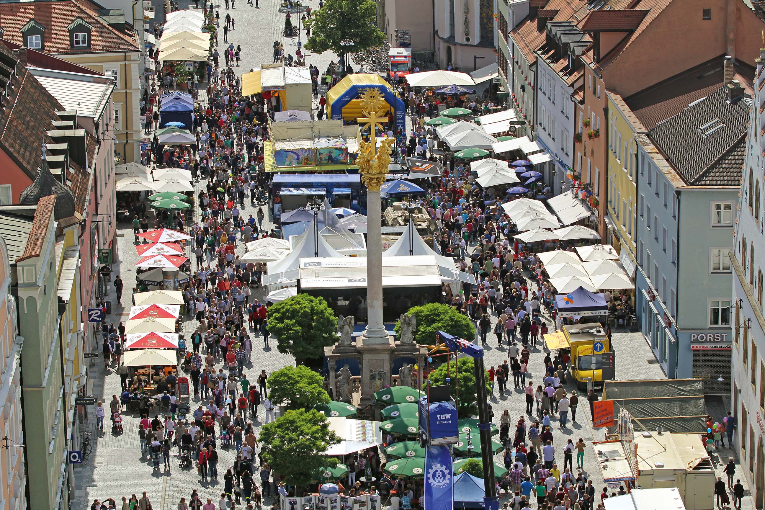Viele Menschen besuchen ein Fest auf dem Straubinger Stadtplatz. Die Sitzplätze werden durch Sonnenschirme angenehm beschattet.