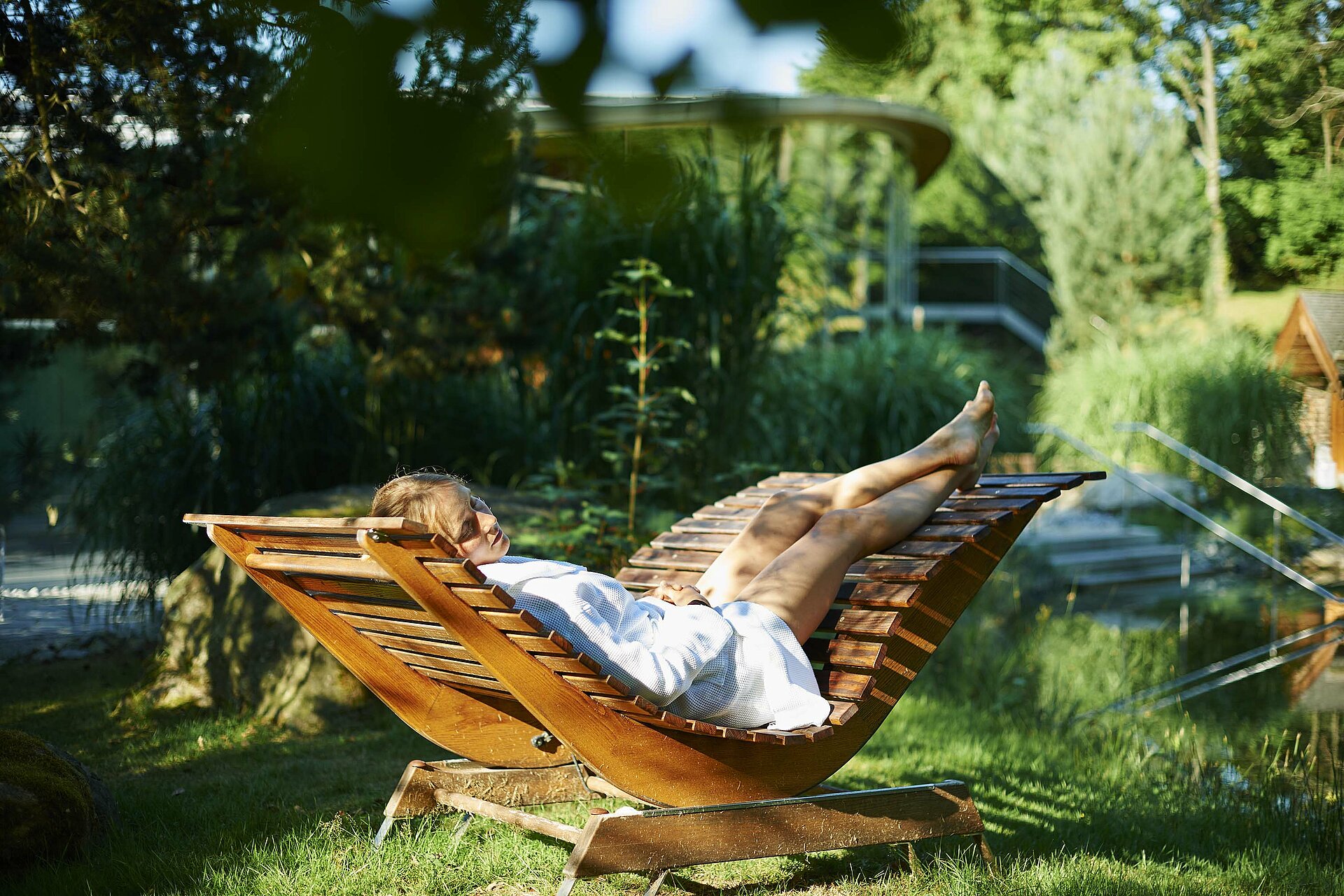 Eine körpergerechte Holzliege steht inmitten einer grünen Oase. Eine junge Frau in weißem Bademantel genießt hier Ruhe und Entspannung pur.