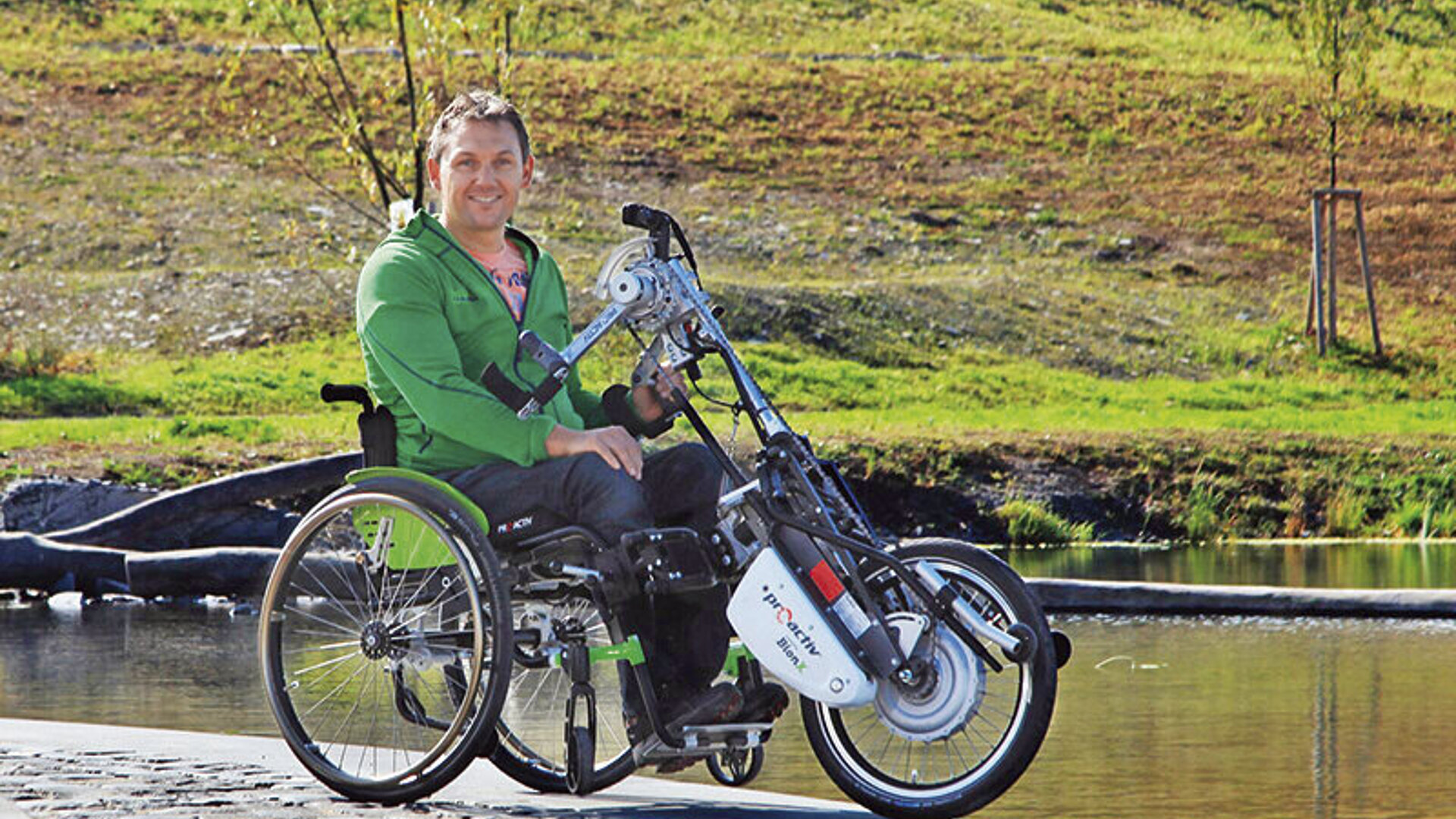 Ein Mann sitzt in einem Rollstuhl mit elektrischem Zuggerät am Rand eines Gewässers und lacht in die Kamera.