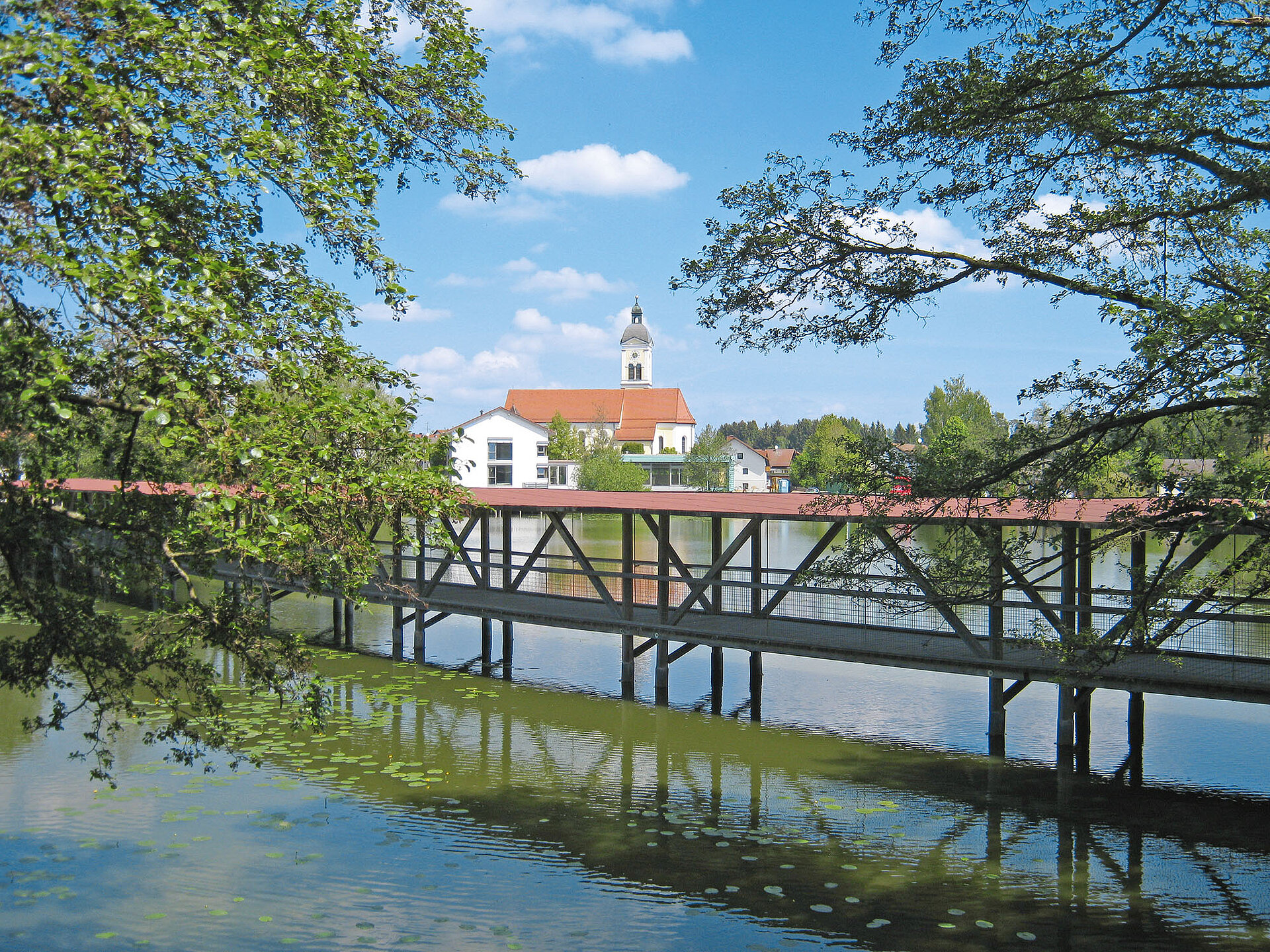 Eine überdachte Holzbrücke für quer über den Beckenweiher und bietet einen schönen Blick das Wasser und auf Wiesenfelden.