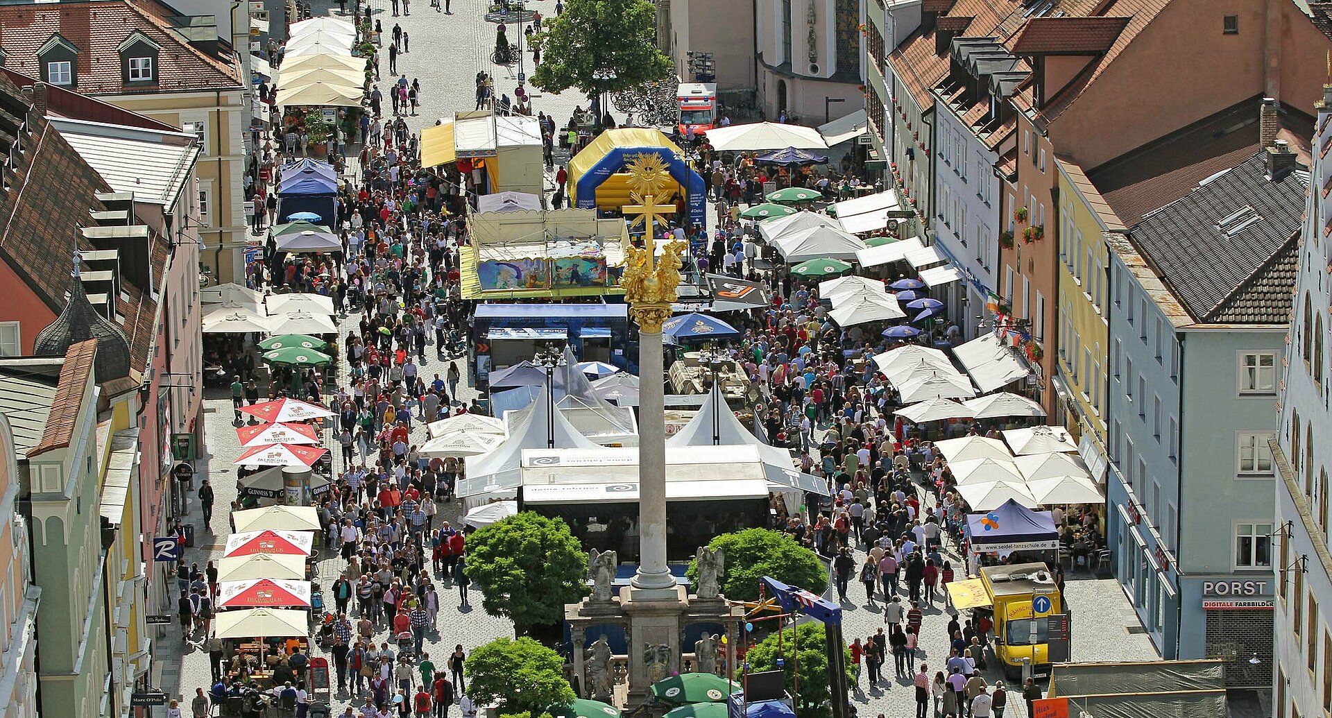 Viele Menschen besuchen ein Fest auf dem Straubinger Stadtplatz. Die Sitzplätze werden durch Sonnenschirme angenehm beschattet.
