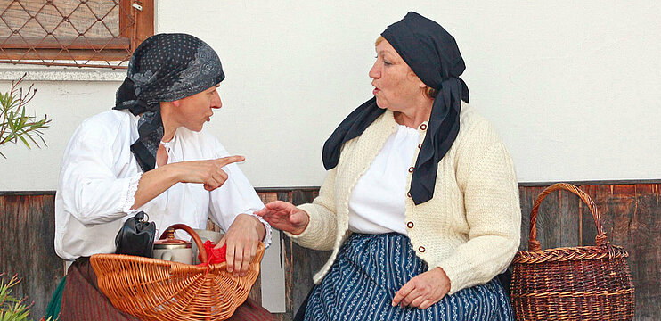 Zwei Frauen sitzen mit Körben auf einer Holzbank vor einem Haus. Beide sind altertümlich gekleidet und die Haaer mit Kopftüchern bedeckt. 