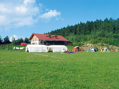 Berghütte und Naturcampingplatz Binder. Auf der großen Wiese vor dem Gebäude befinden sich verschieden große Zelte. Erwachsene und Kinder sitzen davor.