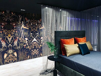 Ein Schlafzimmer mit besonderer Note. Das Bett in Blautönen, dahinter eine feine Gardine und eine Fototapete auf der Eulen, Pilze und verschiende Ornamente dargestellt sind