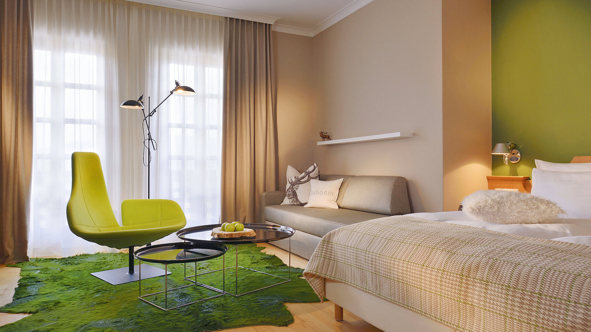 Zimmer mit Bett, Sofa und Sitzbereich, überwiegend gehalten in Grün- und Beigetönen.