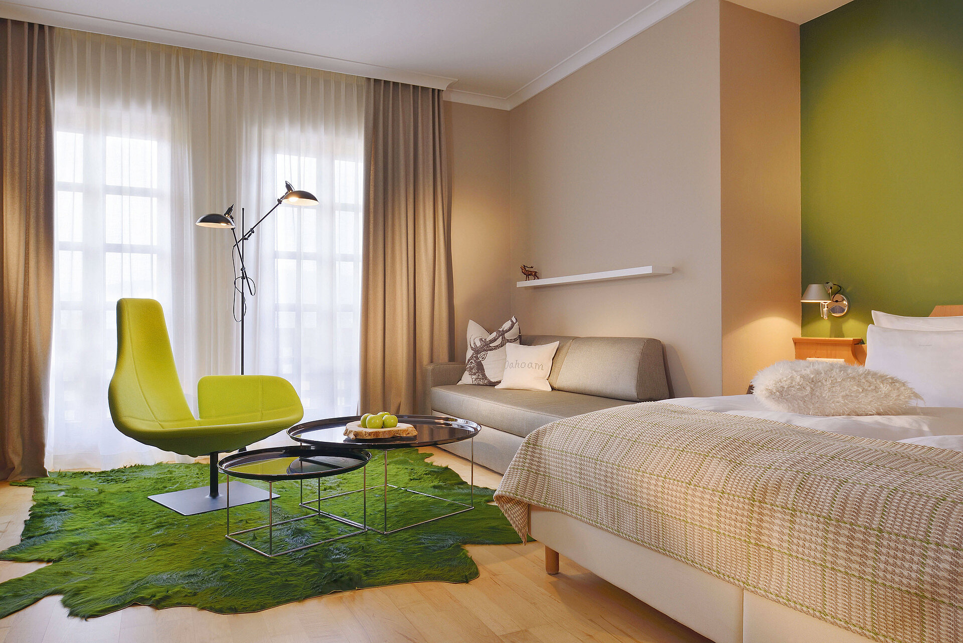 Zimmer mit Bett, Sofa und Sitzbereich, überwiegend gehalten in Grün- und Beigetönen.