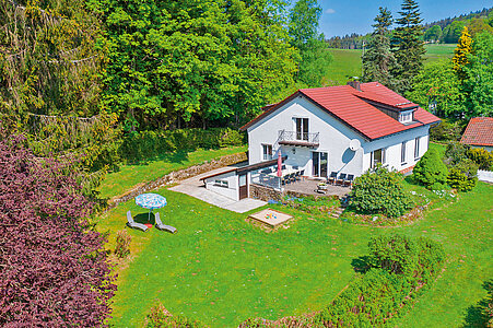 Luftansicht des Ferienhauses am Kapellenweg im Sommer. Das Haus befindet sich in einem großen Gartengrundstück mit Terrasse, Liegwiese, Sandkasten. Hinterm Haus beginnt der Wald.