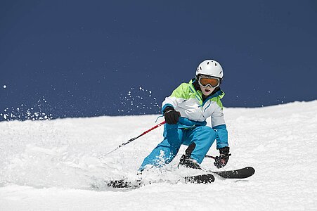 Schnee staubt auf, wenn der junge Skifahrer, perfekt ausgestattet mit Skianzug und Helm, den Skihang rasant und elegant bezwingt.