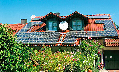 Photovoltaikanlage auf einem Dach mit zwei Dachgauben. Darunter ein Garten, versteckt durch üppig und prachtvoll blühende Pflanzen. 