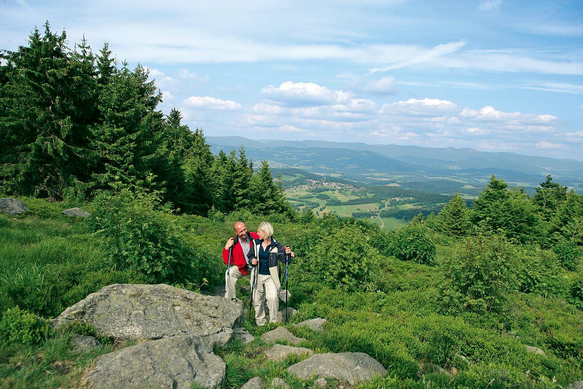 Ein Paar wandert mit Nordic Walking Stöcken von der Nordseite zum Pröllergipfel hoch. Dahinter sieht man einige kleine Bayerwaldorte und die eindrucksvolle Hügelkette des Hinteren Bayerischen Waldes.