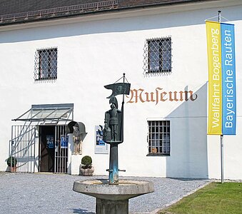 Eingang zum Kreismuseum Bogenberg. Vor dem Museum ein Brunnen, auf dem eine Figur des Heiligen Florian steht. An der Ecke des Museums ist eine gelb blaue Fahne gehißt auf der stehen die Worte Wallfahrt Bogenberg - Bayerische Rauten.