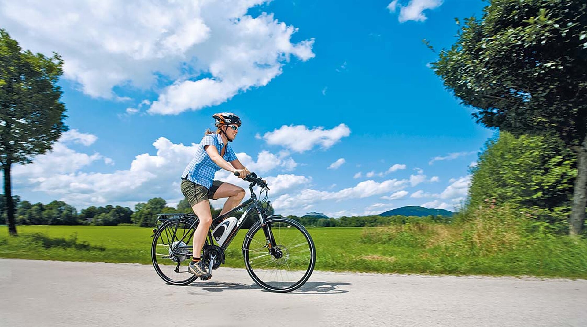 Eine junge Frau genießt die Fahrt mit dem E-Bike. Über ihr der bayerische Himmel weiß und blau.