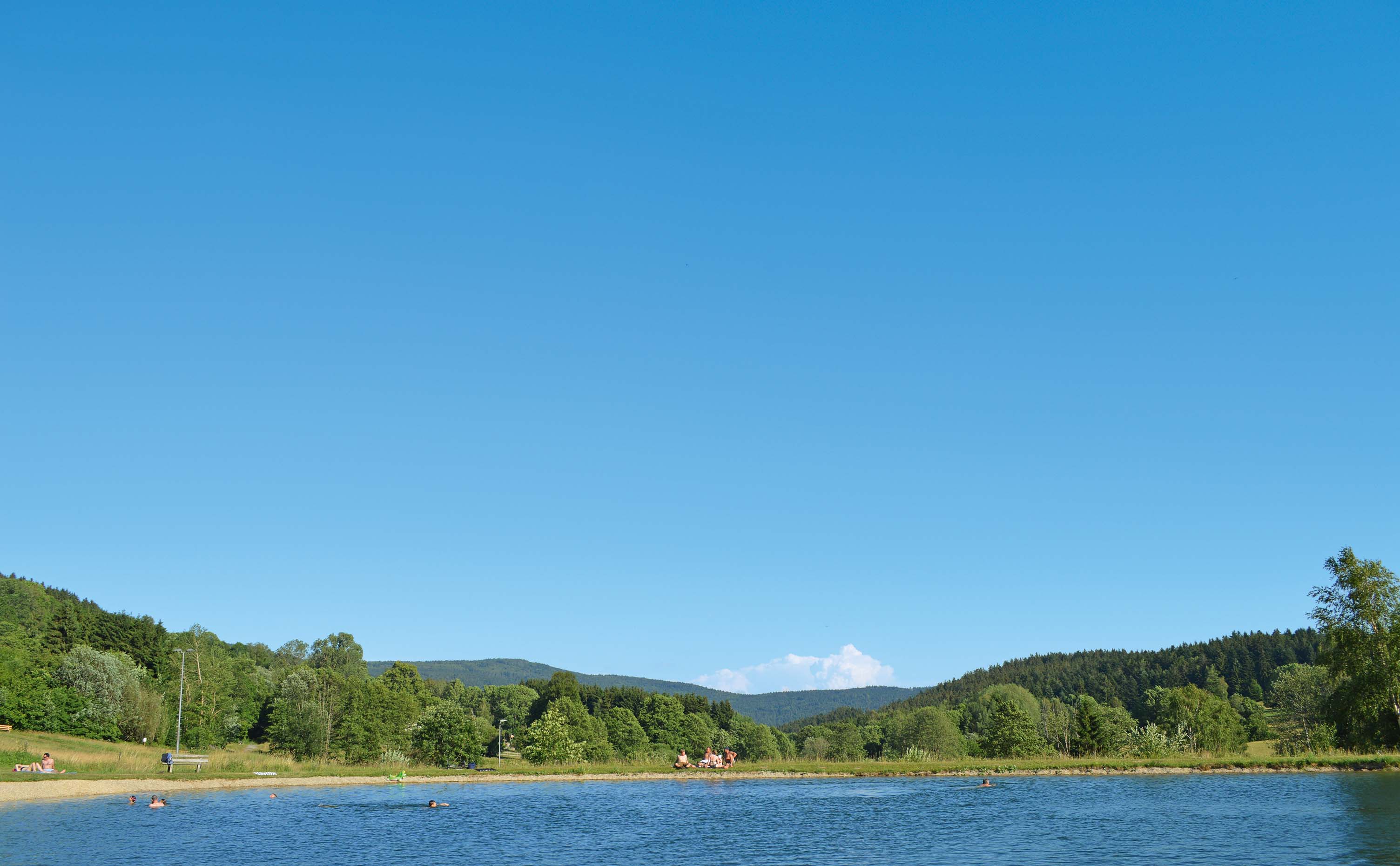 Unter blauem Himmel liegt die Badestelle im Kurpark Sankt Englmar. Im Hintergrund sieht man die Hügellandschaft und den Hirschenstein