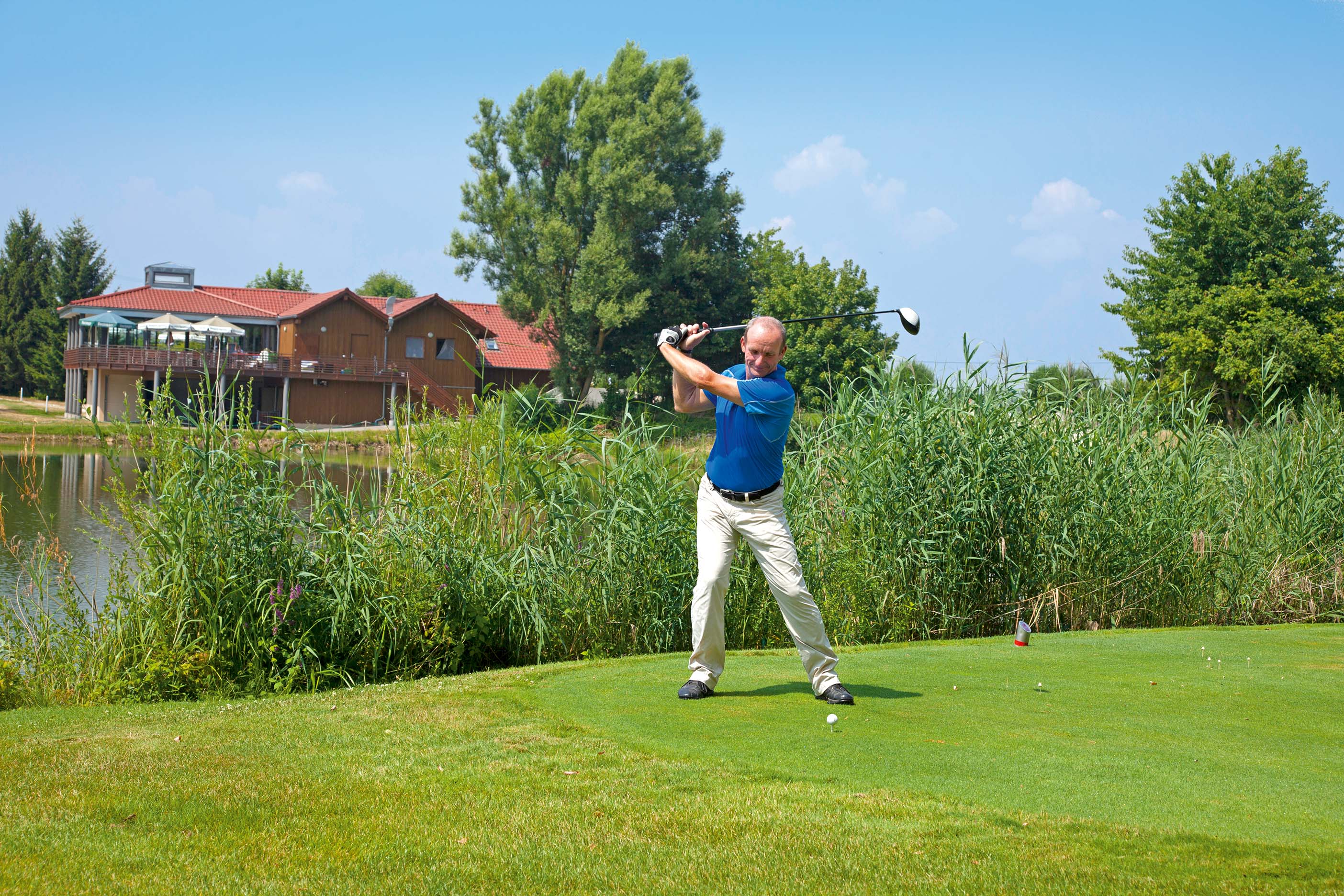 Unter blauem Himmel steht ein Golfspieler in Schlagposition auf grünem Rasen. Hinter Sträuchern verborgen liegt ein Gewässer und ein Gebäude mit Sonnenschirmen auf der Terrasse.