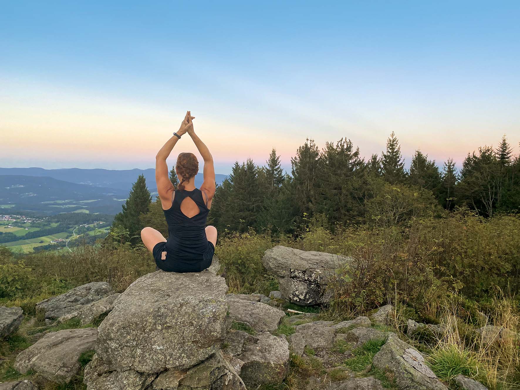 Eine Frau sitzt in Sportbekleidung am Pröllergipfel auf einem Felsen. Während ihr Blick auf dem Tal ruht, vollführt sie eine Yogaübung.