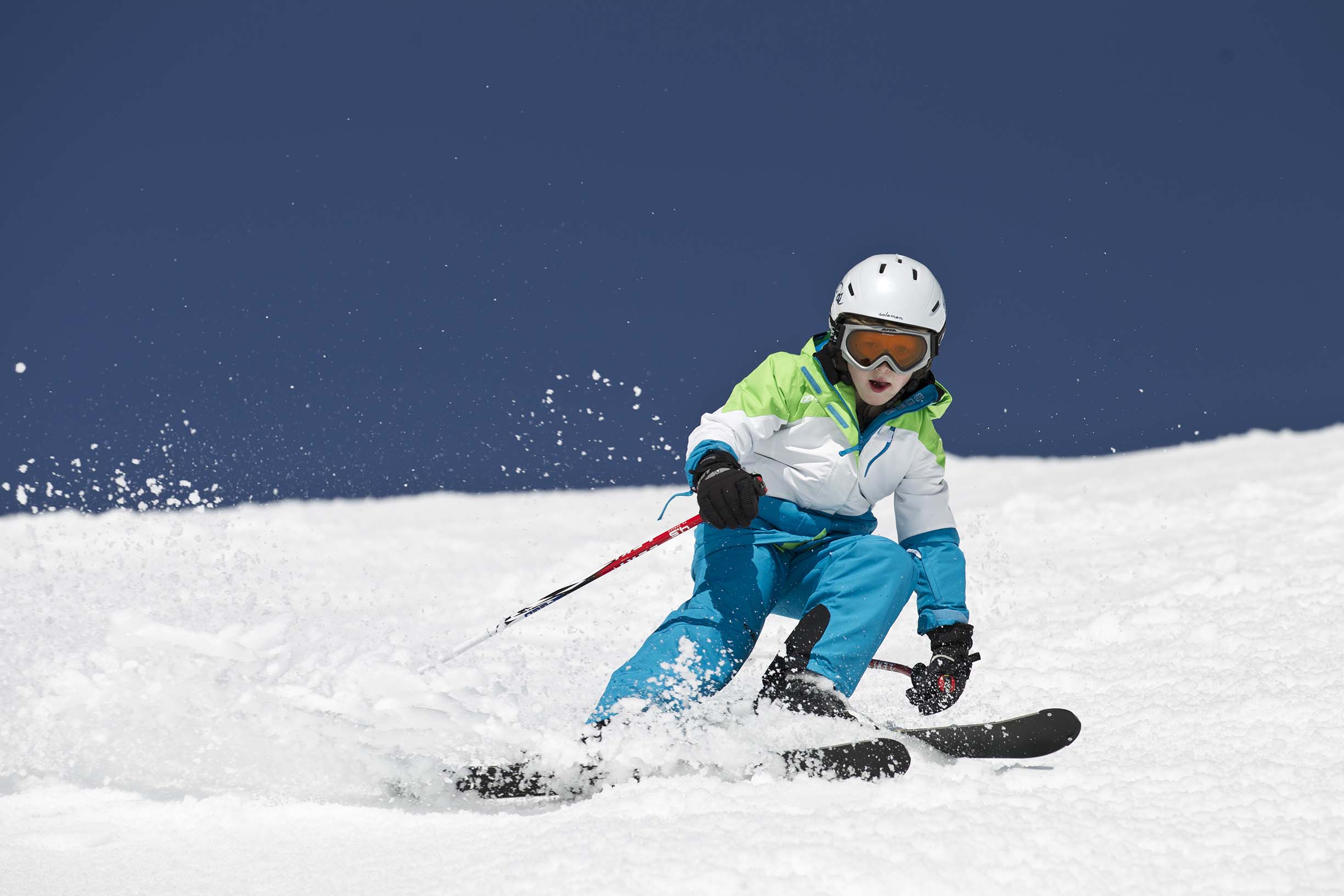 Schnee staubt auf, wenn der junge Skifahrer, perfekt ausgestattet mit Skianzug und Helm, den Skihang rasant und elegant bezwingt.